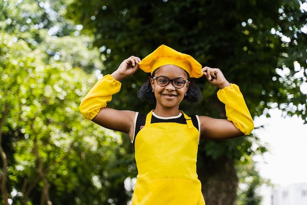 Fille de cuisinier enfant africain noir en chapeau de chefs et uniforme de tablier jaune touchant son chapeau de chefs Publicité créative pour café ou restaurant