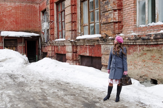 Fille dans un béret rose et un manteau gris-bleu debout dans la cour de la vieille maison