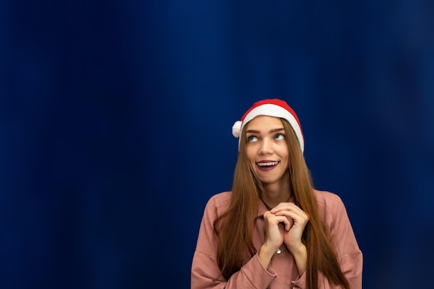 Une fille dans un bonnet de Noel sur un fond bleu isolé lève les yeux vers l'espace de copie Admiration et joie