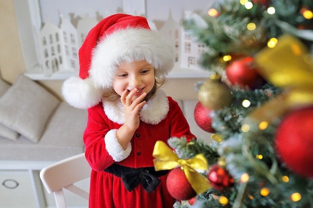 fille dans un bonnet de Noel regarde l'arbre de Noël et sourit en prévision des vacances et d'un cadeau