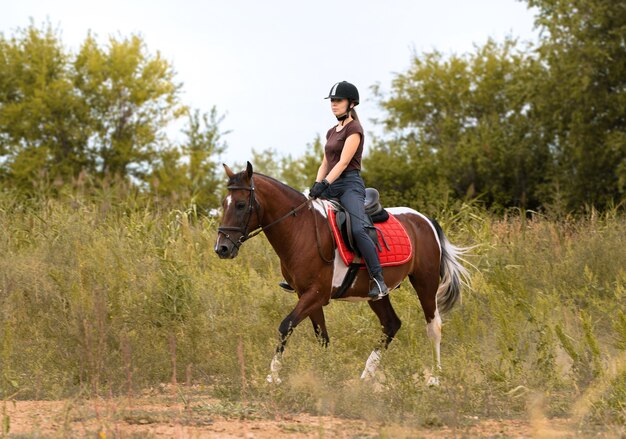 Une fille dans un casque d'équitation monte un cheval skewbald au trot dans un champ parmi les buissons verts vers le photographe