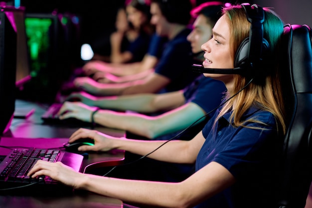 Fille dans le casque avec microphone assis à table et jouer à un jeu vidéo dans un club e-sport