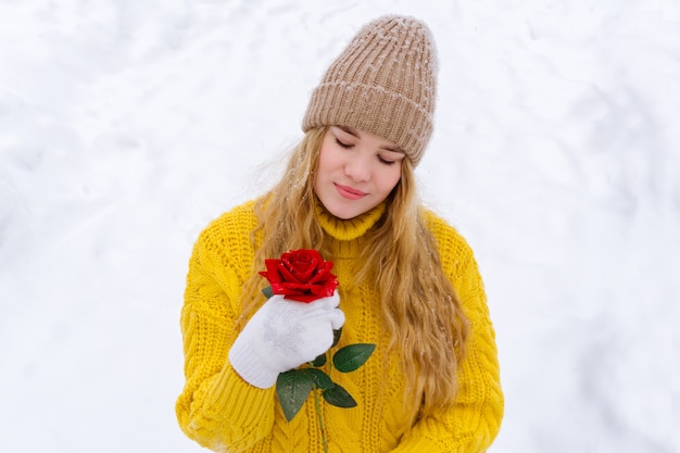 Fille dans un chandail chaud et un chapeau tricoté tient une rose dans ses mains sur un fond enneigé