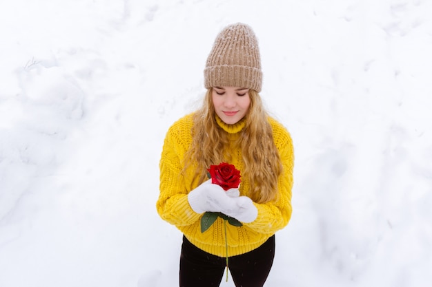 Fille dans un chandail chaud et un chapeau tricoté tient une rose dans ses mains sur un fond enneigé