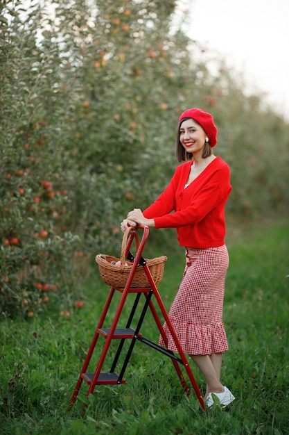 Une fille dans un chandail à jupe rouge et un chapeau se promène dans un verger de pommiers et cherche de bonnes pommes