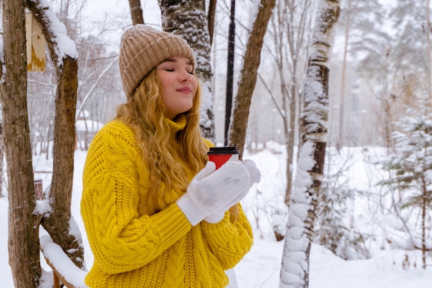 Fille dans un chandail tenant une tasse de papier avec une boisson chaude dans un parc d'hiver