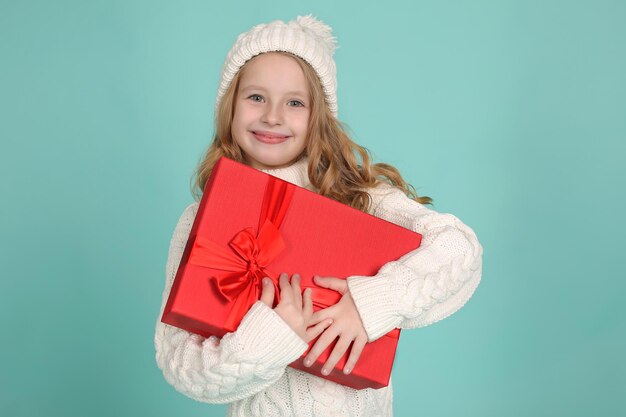 fille dans un chapeau blanc tricoté et un pull blanc tricoté sur fond bleu étreint une boîte cadeau rouge