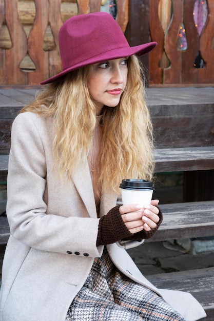 Une fille dans un chapeau boit du café assis sur des marches en bois à l'automne