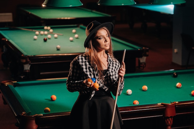 Une fille dans un chapeau dans un club de billard avec une queue dans ses mains. Jeu de billard.