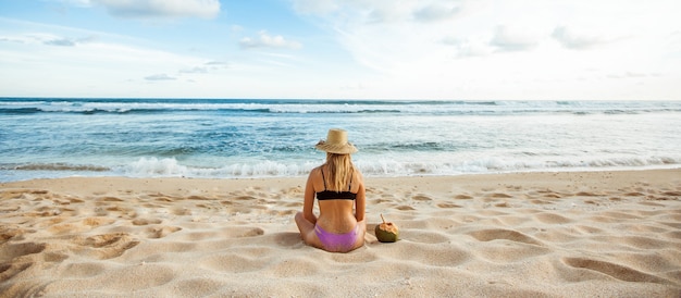 Une fille dans un chapeau est assis sur le sable blanc avec son dos à la caméra et regarde l'océan à côté est un panorama de la bannière de noix de coco