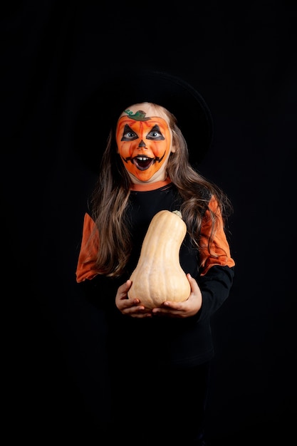 Une fille dans un chapeau noir et avec du maquillage de citrouille sur son visage tient une citrouille dans ses mains et célèbre Halloween, isolé