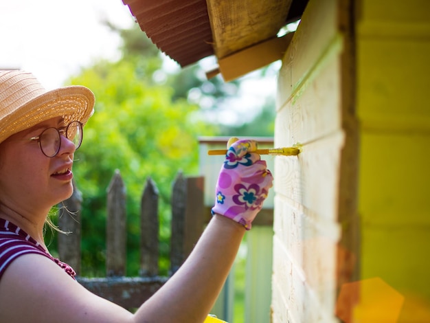 Une fille dans un chapeau peint un mur en bois avec de la peinture jaune avec un pinceau une mère dans le pays peint une maison pour un enfant