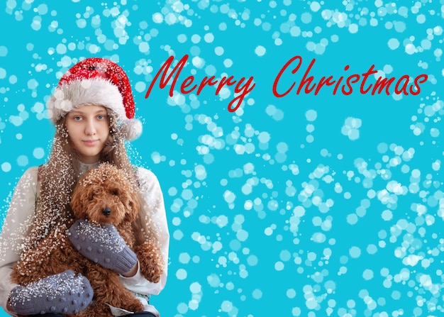 Une fille dans un chapeau de Père NoëlUne fille dans un chapeau de Père Noël tient un caniche dans la neige en mitaines dans ses bras une carte de joyeux Noël tient un caniche dans ses bras