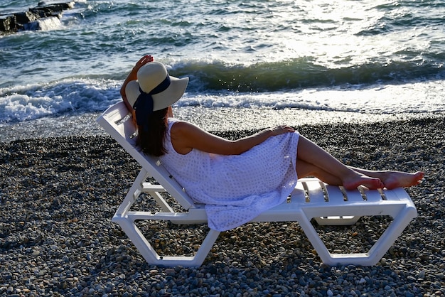 une fille dans un chapeau et une robe blanche d'été est allongée sur la plage et regarde dans la distance