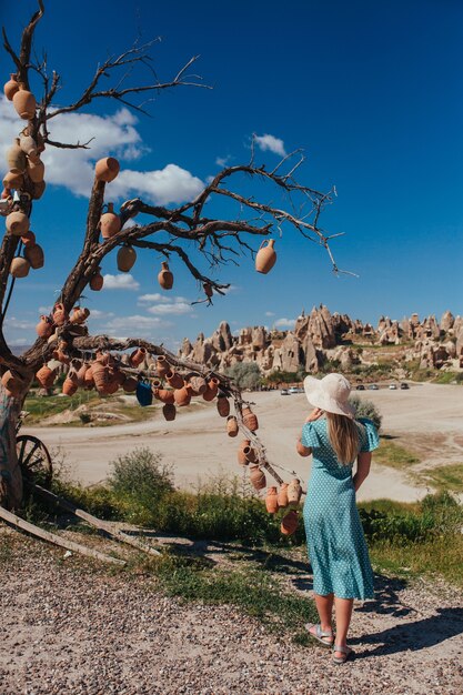 Une fille dans un chapeau et une robe rose se tient près d'un arbre accroché avec des plats en céramique en capadocia