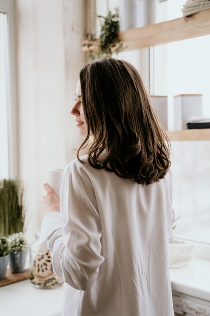 Fille dans une chemise blanche boit du café le matin dans une cuisine.