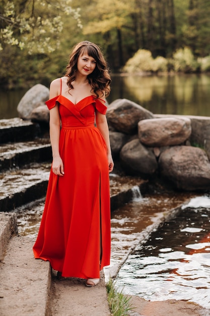 Fille dans une longue robe rouge près du lac dans le parc au coucher du soleil.