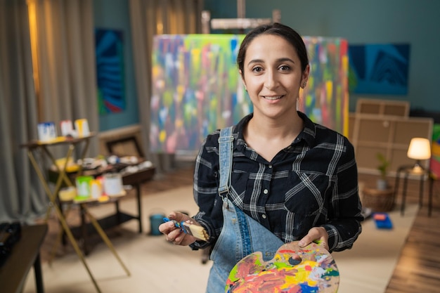 Fille dans la main sale de la peinture tient le pinceau et la palette en bois pour les peintures sourire sur son visage rire