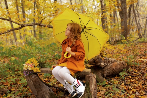 fille dans un manteau beige dans un automne urbain est assise sur un banc avec un parapluie jaune dans ses mains