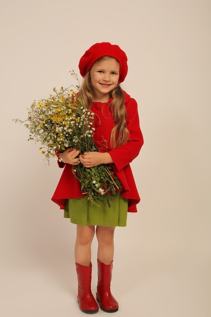 une fille dans un manteau rouge, des bottes et une robe verte et un béret rouge tient des fleurs sauvages dans ses mains