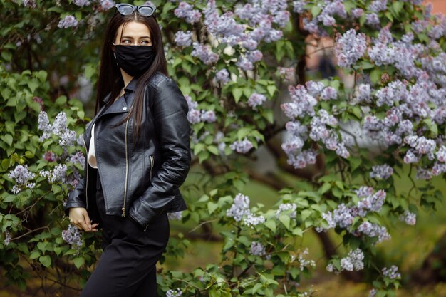 Fille dans un masque médical sur le fond d'un lilas en fleurs dans le parc. Masque noir. Protection contre les virus, la grippe