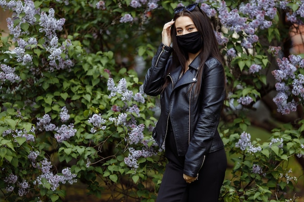 Fille dans un masque médical sur les lilas en fleurs. Masque noir. Protection contre les virus, la grippe. Protection contre le coronavirus. épidémie de coronavirus. l'arôme d'un arbre dans le jardin Allergie printanière.