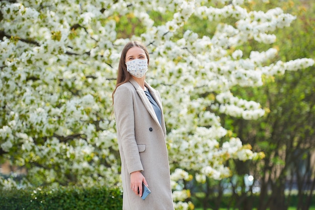 Une fille dans un masque médical pour éviter la propagation du coronavirus est titulaire d'un smartphone dans le parc. Une femme dans un masque facial contre COVID-19 maintient une distance sociale dans le jardin entre les arbres en fleurs.