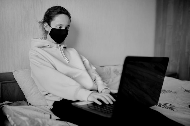 Fille dans un masque noir travaille sur un ordinateur portable à la maison en isolement photo noir et blanc