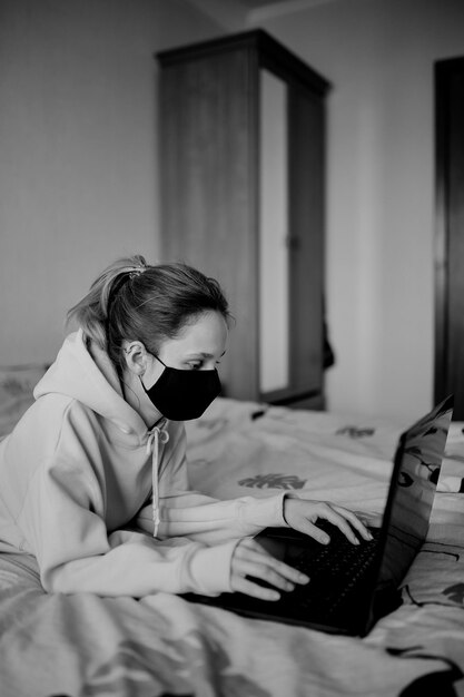 Fille dans un masque noir travaille sur un ordinateur portable à la maison en isolement photo noir et blanc