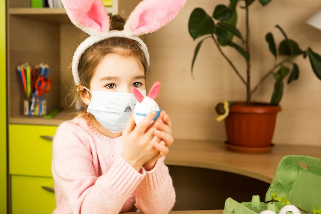 Fille dans les oreilles de lièvre fait un lapin de Pâques dans un masque médical à partir d'un œuf et de la pâte à modeler.