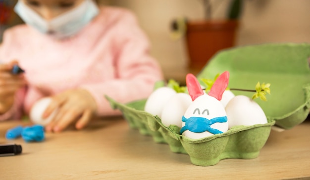 Fille dans les oreilles de lièvre fait un lapin de Pâques dans un masque médical à partir d'un œuf et de la pâte à modeler.