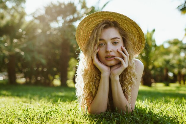 Une fille dans le parc est allongée sur l'herbe, soutenant ses joues avec ses mains et regardant la caméra de manière romantique