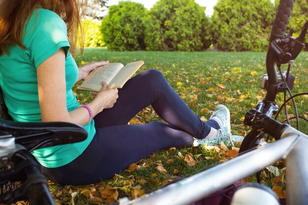 Une fille dans le parc est assise sous un arbre et lit un livre. à côté il y a un vélo et un sac à dos