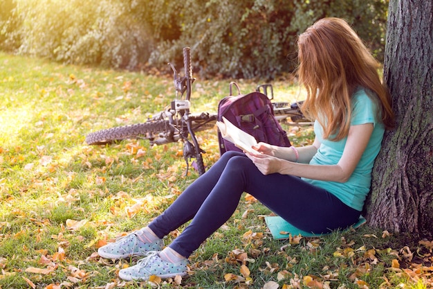 Une fille dans le parc est assise sous un arbre et lit un livre. à côté il y a un vélo et un sac à dos