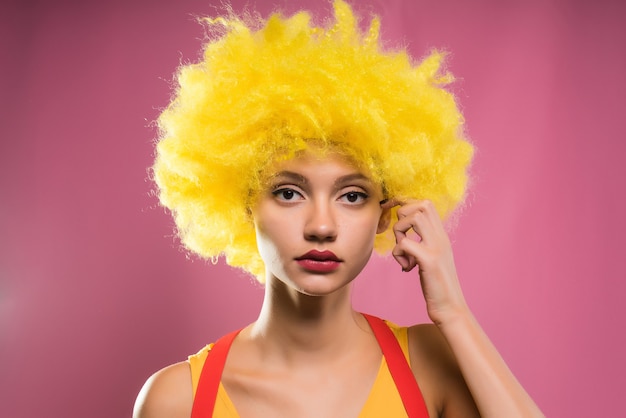 Fille dans une perruque jaune vif regarde la caméra, isolée sur fond rose