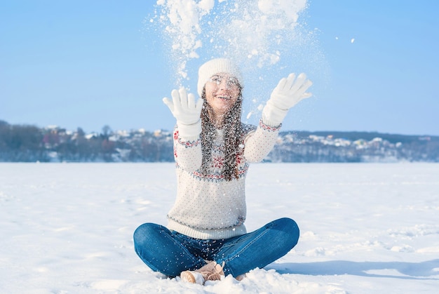 Fille dans un pull d'hiver blanc S'assoit avec les jambes croisées dans la neige Jette la neige vers le haut