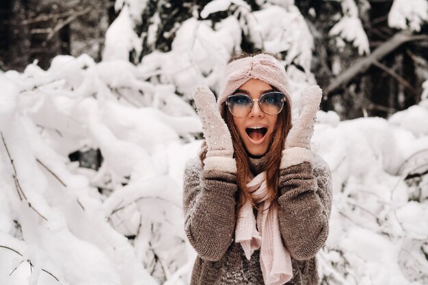 Une fille dans un pull et des mitaines en hiver tient sa tête avec ses mains dans une forêt enneigée