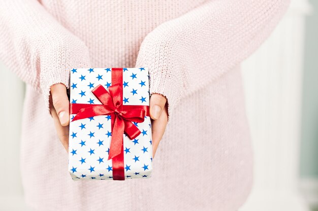 Une fille dans un pull rose tient dans ses mains un cadeau avec des étoiles bleues et un ruban rouge. Sur un fond clair. Concept sur le thème des vacances.