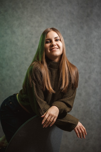 Fille dans un pull vert et un jean sur une chaise grise et souriant