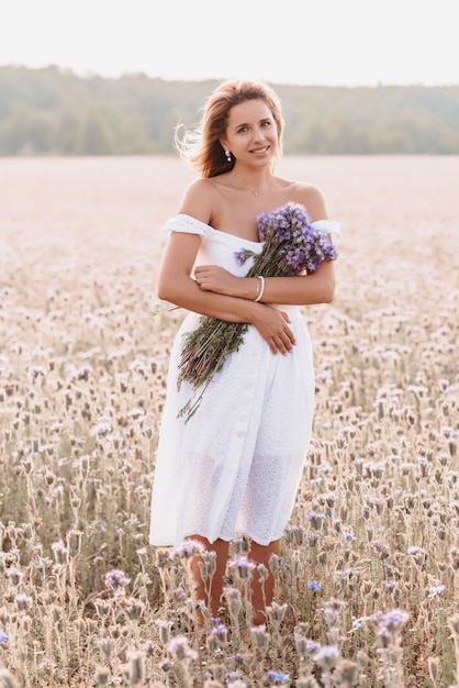 Fille dans une robe blanche avec un bouquet de fleurs violettes dans un champ dans la nature en été