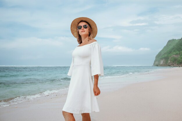 Fille dans une robe blanche se promène sur une plage de sable blanc dans un chapeau et des lunettes de soleil