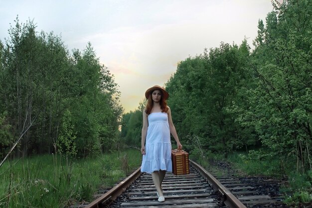 Fille dans une robe d'été blanche et une valise en osier marchant sur des rails