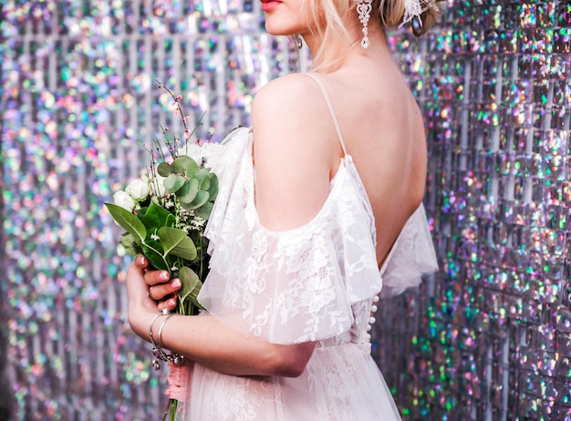 Une fille dans une robe de mariée blanche avec des épaules nues maquillage une coiffure chignon pose sur fond de paillettes La fille se tient dos à la caméra