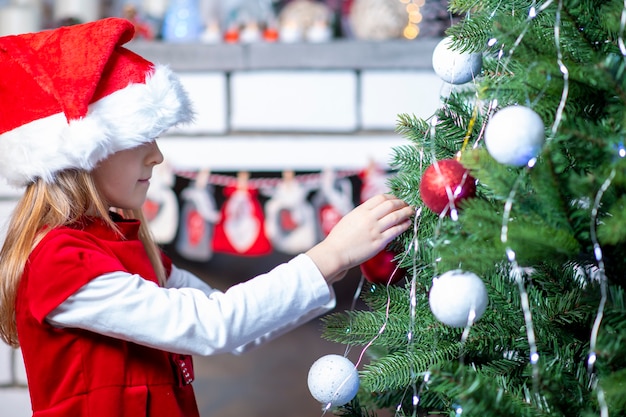 Une fille dans une robe rouge et un chapeau de père Noël près de la cheminée s'accroche à un jouet sur l'arbre de Noël