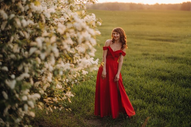 Une fille dans une robe rouge avec des lèvres rouges se tient à côté d'un grand arbre fleuri blanc au coucher du soleil.
