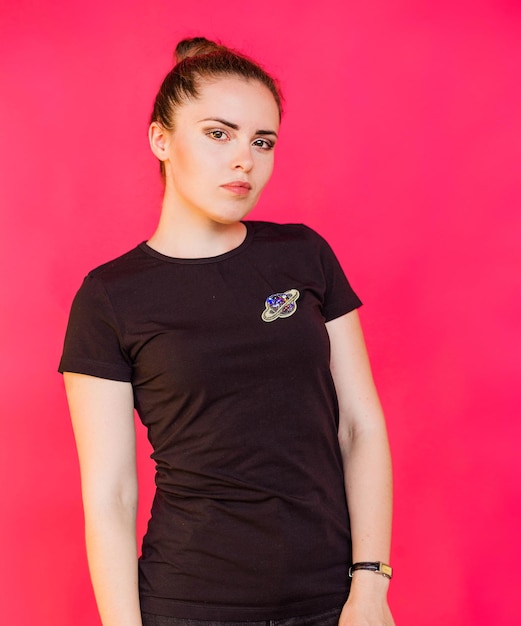 Une fille dans un T-shirt noir avec une broche en forme de planète avec des anneaux pose sur un fond fuchsia