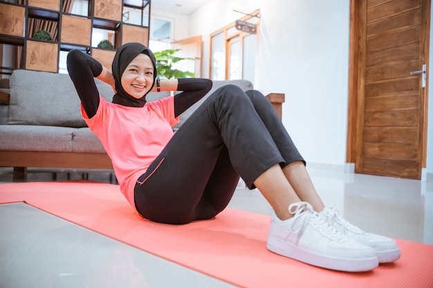 Une fille dans une tenue de gym hijab sourit tout en faisant un sit-up sur un tapis sur le sol dans la maison