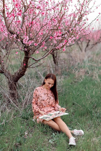 Une fille dans un verger de pêchers en fleurs se repose sous un arbre assis et dessinant