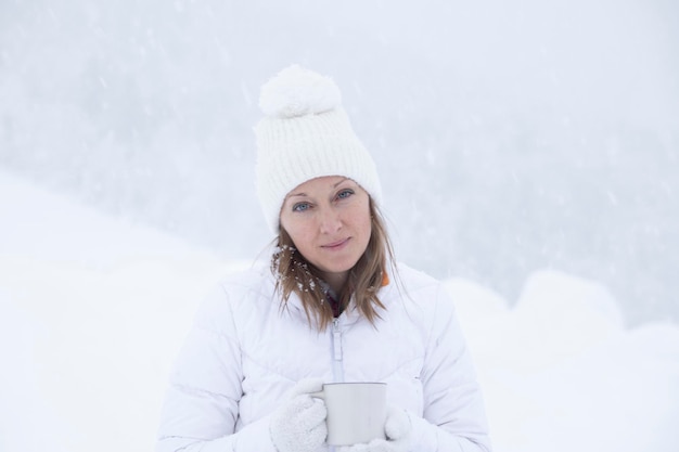 Une fille dans une veste blanche et un chapeau blanc tient une tasse de thé dans ses mains pendant une chute de neige sur fond de belles congères