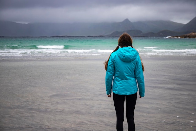 une fille dans une veste bleue admire le paysage sombre sur une plage des îles lofoten en norvège